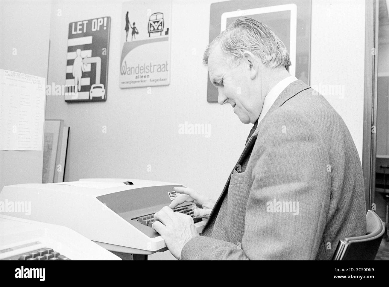 Mr. Holtslag, Haarlem police, police, Haarlem, Nederland, 04-12-1979 Whizgle News, Dutch Desk, pays-Bas, 1950-2000 Un homme en costume se concentre sur la frappe d'une machine à écrire électronique vintage, entouré d'affiches de style rétro sur le mur qui présentent divers signes et messages. Son attitude attentive suggère un engagement envers la tâche à accomplir. Banque D'Images