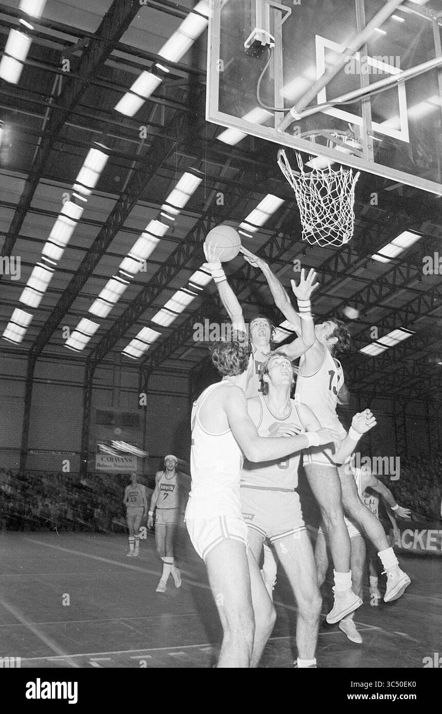 Match de basket Whizgle News, Dutch Desk, pays-Bas, 1950-2000 Un match de basket-ball tendu se déroule alors qu'un joueur se lance dans un layup tout en faisant face à une défense intense de deux adversaires, le tout sous les lumières brillantes de l'arène de style industriel. Les coéquipiers et les spectateurs sont visibles en arrière-plan, ajoutant à l'excitation du moment. Banque D'Images