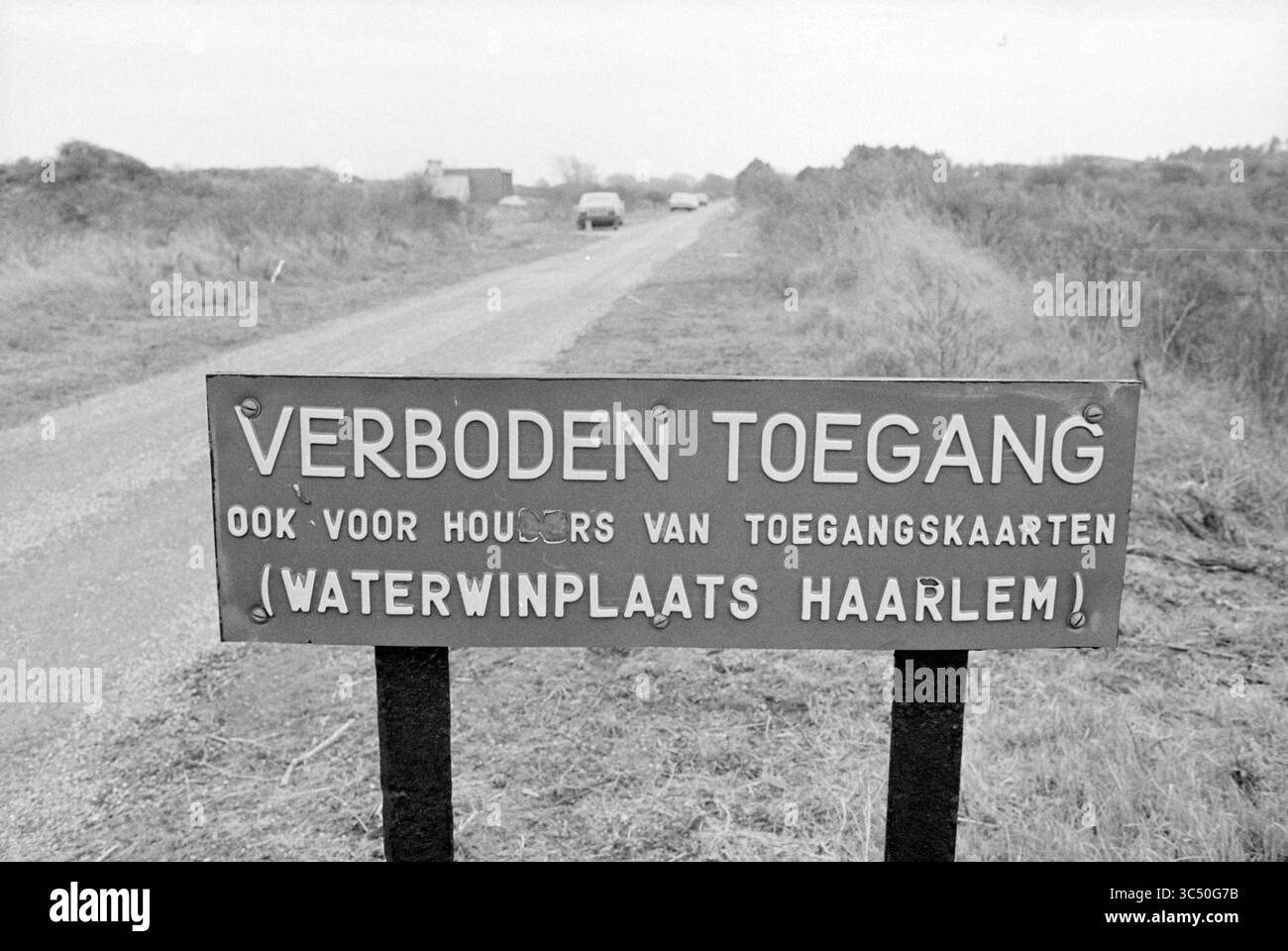 Excursion Water Company Haarlem, Water Supply, Haarlem, pays-Bas, 07-05-1979 Whizgle News, Dutch Desk, pays-Bas, 1950-2000 Une pancarte altérée se trouve au bord d'un chemin de gravier, affichant les mots « Verboden Toegang » qui se traduit par « accès non autorisé ». Il indique des restrictions, en particulier pour les personnes sans laissez-passer d'entrée, liées à une installation d'extraction d'eau à Haarlem. La région environnante présente une végétation clairsemée et une vue lointaine des véhicules le long de la route. Banque D'Images