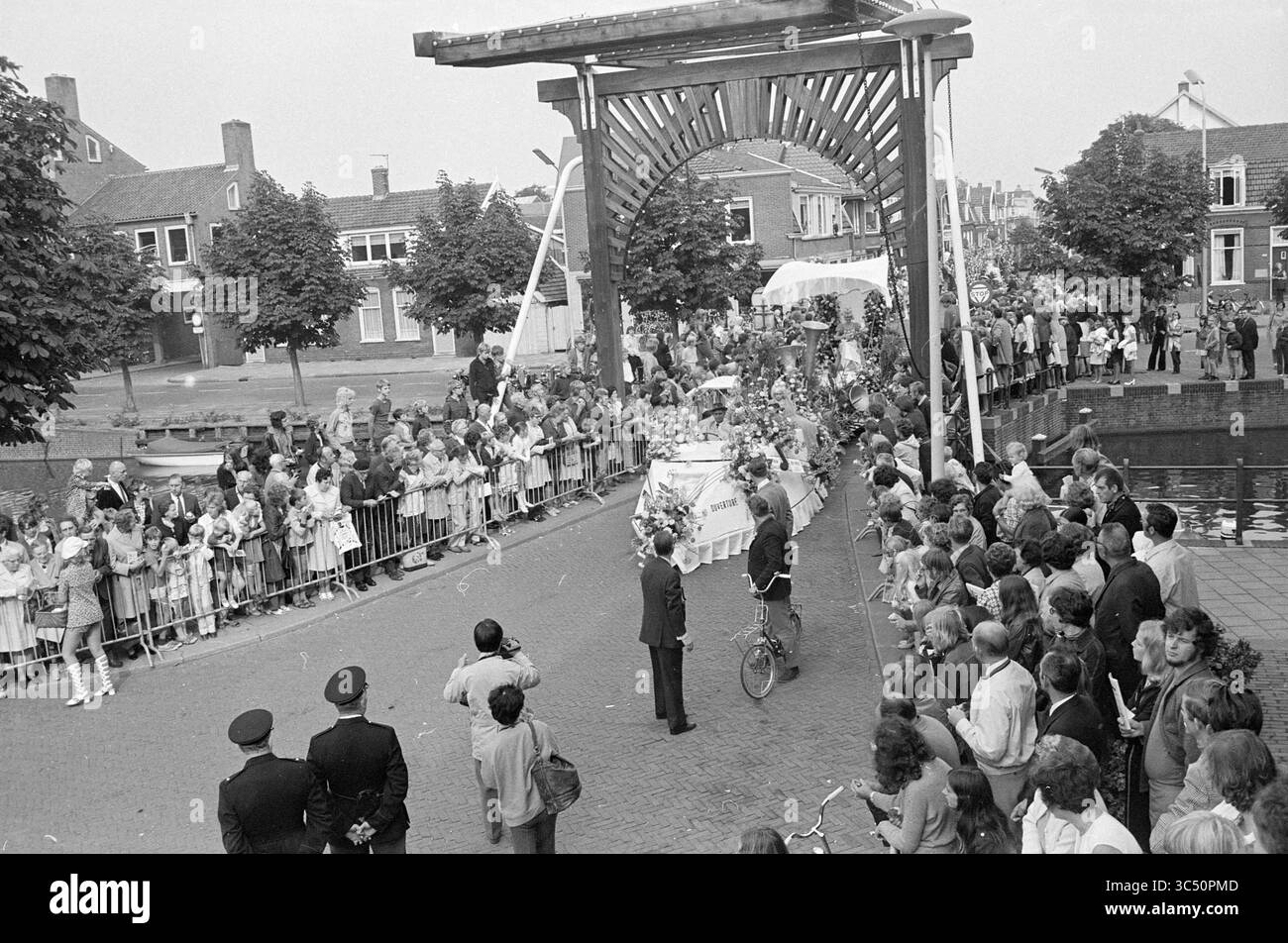 Flower Parade Aalsmeer, Flower Parade, exposition de fleurs, 04-09-1971 Whizgle News, Dutch Desk, pays-Bas, 1950-2000 Une foule nombreuse se rassemble le long d'un canal, bordé d'arbres et de bâtiments, pour assister à une procession festive. Les gens affichent un mélange d'excitation et de curiosité alors qu'un véhicule décoré passe lentement sous un pont orné. Les officiers en uniforme maintiennent l'ordre pendant que les enfants et les adultes prennent des photos et font la vague. Banque D'Images