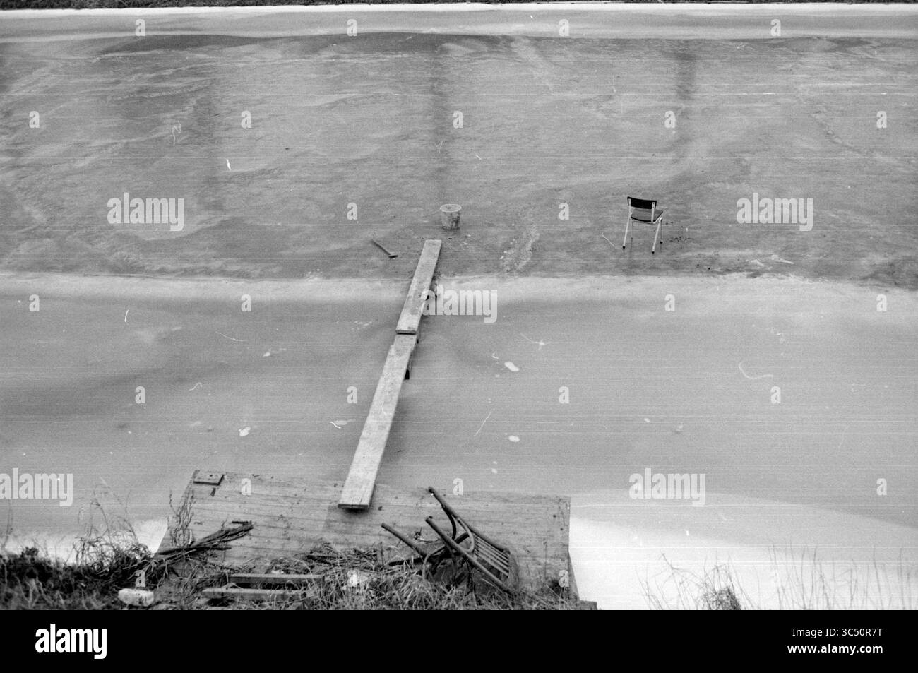 Planche avec chaise sur glace Hoofdvaart, glace, patinoire, hockey sur glace, 19-01-1987 Whizgle News, Dutch Desk, pays-Bas, 1950-2000 Une chaise solitaire se trouve sur la surface glacée d'un plan d'eau gelé, à côté d'une planche de bois s'étendant d'un quai usé. À proximité, des débris dispersés suggèrent un espace oublié, où la nature et la négligence s’entrelacent. Banque D'Images