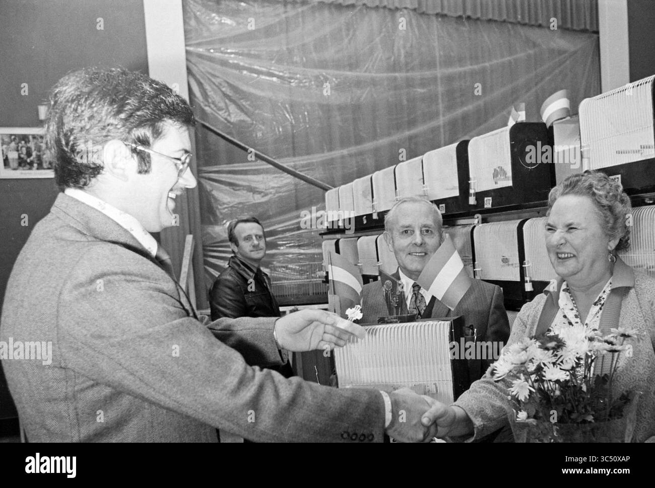 Lauréat de l'exposition d'oiseaux IJmuiden, oiseaux, 04-12-1975 Whizgle News, Dutch Desk, pays-Bas, 1950-2000 Un homme serre joyeusement la main d'une femme, tous deux souriants, tandis qu'un homme plus âgé distingué regarde. En arrière-plan, plusieurs boîtes ou appareils sont soigneusement disposés, faisant allusion à un rassemblement ou à un événement. L'atmosphère transmet un sentiment de célébration et de camaraderie. Banque D'Images