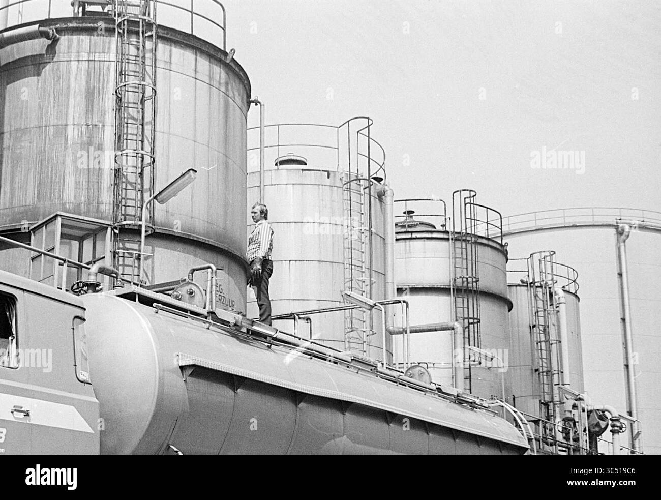 Usine d'acide nitrique Hoogovens Mekog., Hoogovens, 10-07-1974 Whizgle News, Dutch Desk, pays-Bas, 1950-2000 Une personne se tient au sommet d'un grand camion-citerne, surplombant une série de réservoirs de stockage industriels avec des échelles métalliques et des tuyauteries. La scène transmet un sens de l'échelle et de l'activité industrielle. Banque D'Images