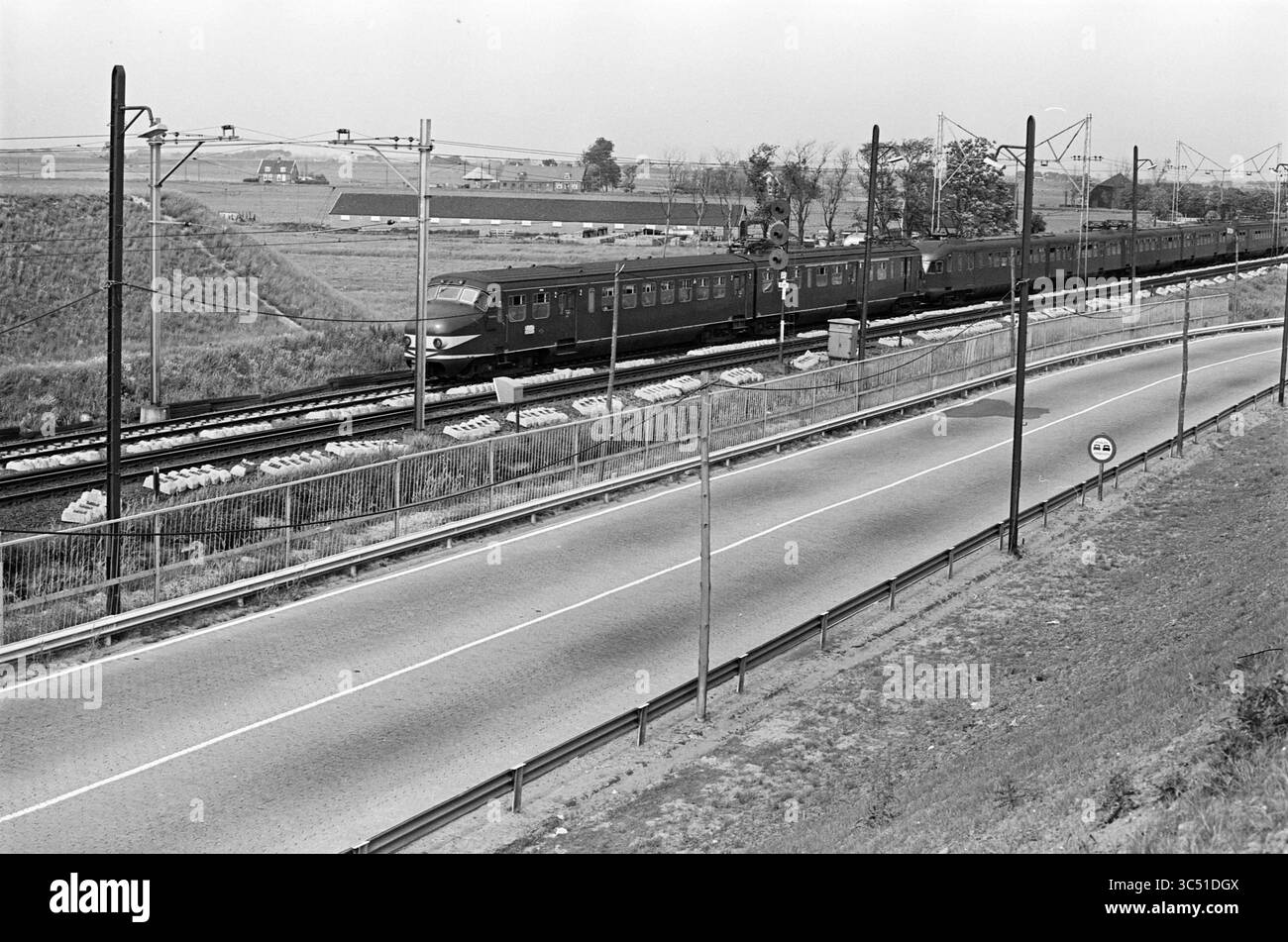 Train et bus Haarlem - Amsterdam, Noord Zuid Hollandse Vervoersmaatschappij N.V., NZ, 11-06-1964 Whizgle News, Dutch Desk, pays-Bas, 1950-2000 Un train épuré glisse le long de voies bordées de champs herbeux et de structures lointaines, tandis qu'une route parallèle longe le train, marquée par un panneau de signalisation. La scène capture un moment tranquille mais dynamique de transport dans un cadre rural. Banque D'Images
