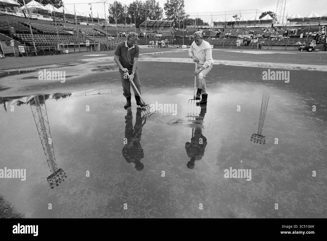 Rain, Pim Mulier Stadium, 11-07-1992 Whizgle News, Dutch Desk, pays-Bas, 1950-2000 deux personnes travaillent avec diligence sur un champ humide et couvert de flaques d'eau, en utilisant des fourches pour briser la surface pendant qu'elles préparent la zone, avec leurs reflets visibles dans l'eau peu profonde. L'arrière-plan présente des stands et des structures vides, suggérant un lieu sportif. Banque D'Images
