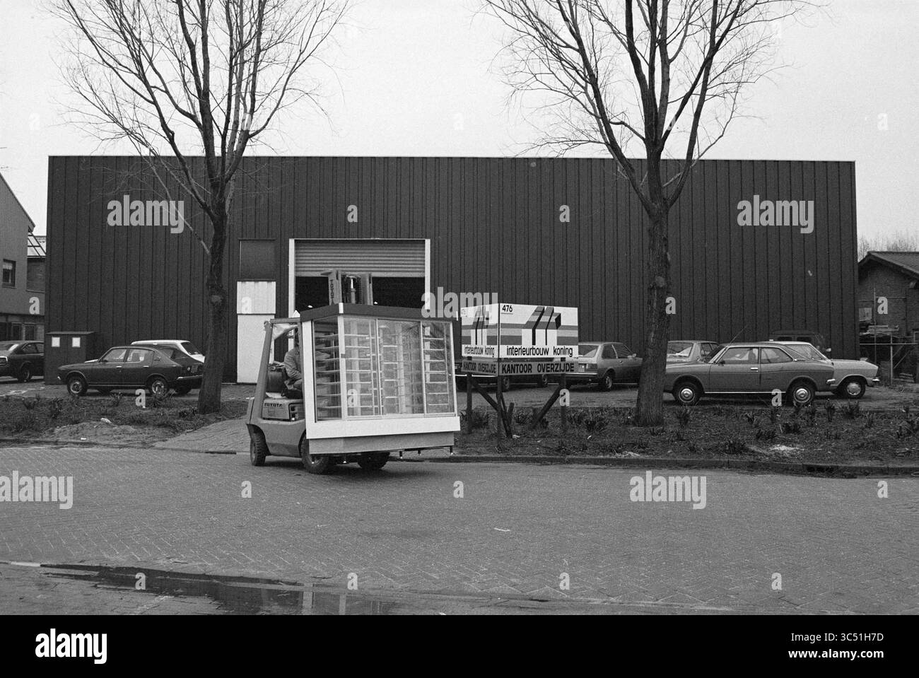 Rapport sur la construction intérieure de Koning à Vijfhuizen, construction, chantiers, intérieur, Vijfhuizen, 23-01-1980 Whizgle News, Dutch Desk, pays-Bas, 1950-2000 Un petit véhicule de livraison transporte des piles de produits, s'éloignant d'un grand bâtiment industriel. Autour de la structure se trouvent plusieurs voitures garées et des arbres arides, contribuant à l'atmosphère animée mais tamisée de la région. Un panneau à proximité indique le nom de l'entreprise et les services offerts. Banque D'Images