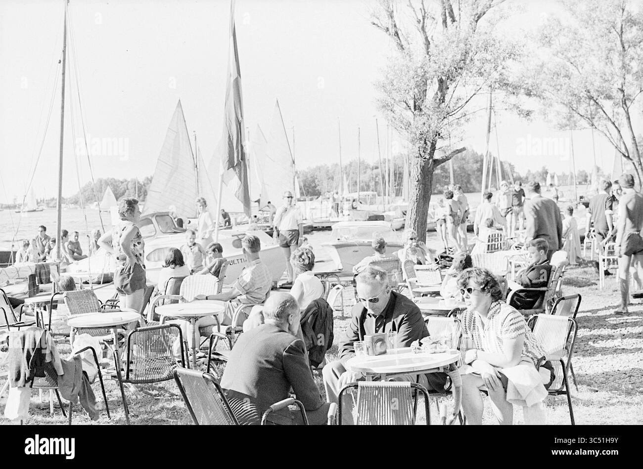 Kaageiland, Summer Pictures, Busyness, 21-07-1963 Whizgle News, Dutch Desk, les pays-Bas, 1950-2000 Une scène animée au bord de l'eau avec des gens prenant un verre à des tables en plein air, entouré de voiliers et d'activités au bord de l'eau. Certaines personnes s'engagent dans la conversation tandis que d'autres prennent le soleil, créant une atmosphère vibrante remplie d'interactions sociales et de loisirs. Les arbres bordent l'arrière-plan, ajoutant une touche de nature à l'ambiance animée. Banque D'Images
