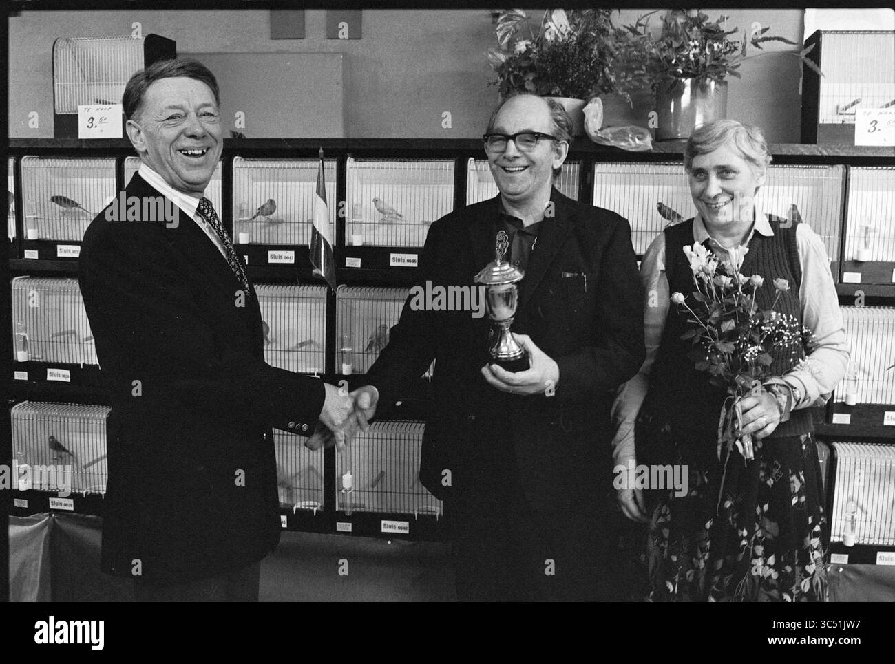 Remise du trophée Damiate à l'association Velsen Bird, cérémonie de remise des prix, Birds, Velsen, 02-12-1979 Whizgle News, Dutch Desk, pays-Bas, 1950-2000 Un homme serre la main à un autre homme tenant un trophée, tandis qu'une femme à côté d'eux présente un bouquet de fleurs. Ils sont entourés de cages remplies d'oiseaux, suggérant une célébration ou une cérémonie de remise de prix dans un spectacle d'oiseaux ou un événement similaire. Banque D'Images