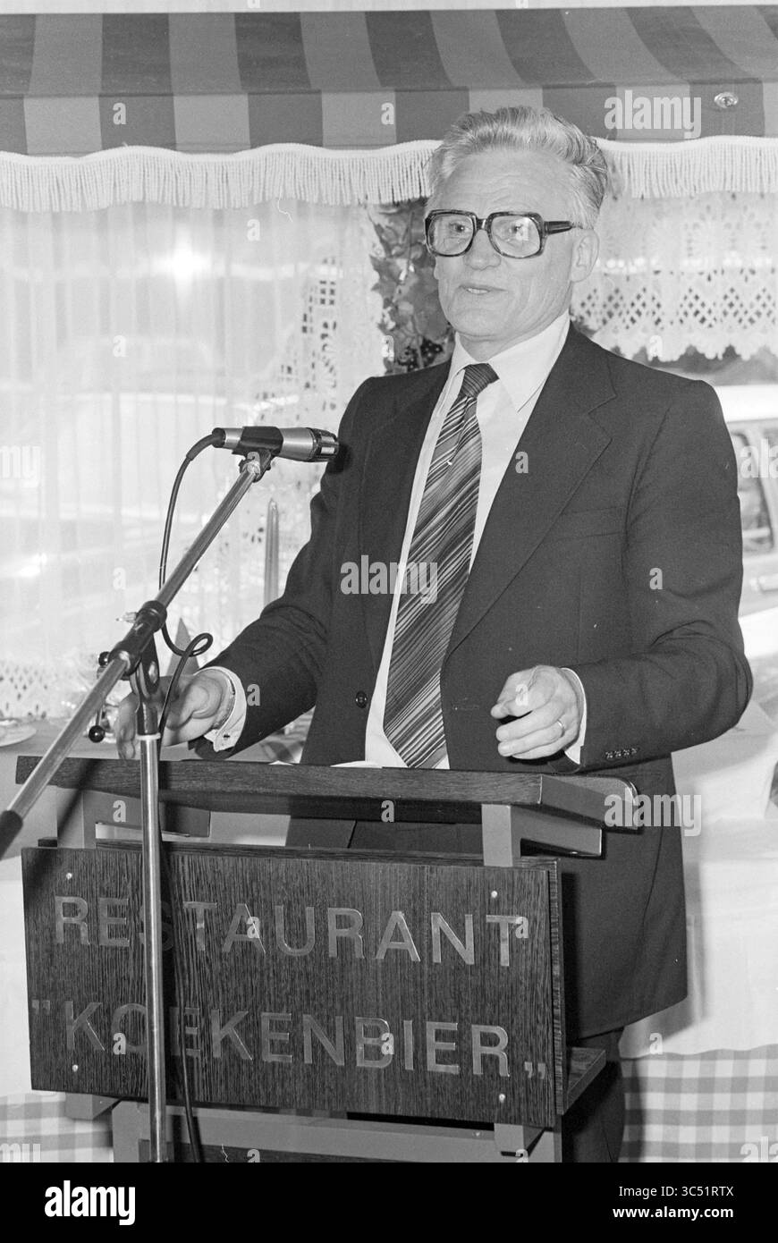 Portrait d'un homme Whizgle News, Dutch Desk, pays-Bas, 1950-2000 Un homme se tient debout sur un podium s'adressant à un public, portant un costume et une cravate, avec des lunettes et un comportement confiant. La toile de fond présente des éléments décoratifs, faisant allusion à un événement formel ou communautaire, peut-être lié à un restaurant. Banque D'Images Portrait d'un homme Whizgle News, Dutch Desk, pays-Bas, 1950-2000 Un homme se tient debout sur un podium s'adressant à un public, portant un costume et une cravate, avec des lunettes et un comportement confiant. La toile de fond présente des éléments décoratifs, faisant allusion à un événement formel ou communautaire, peut-être lié à un restaurant. Banque D'Images