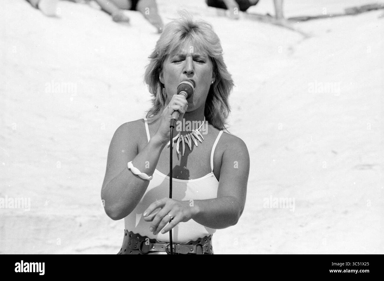 Anita Meyer at riche Zandvoort, musique, plages, photos d'été, Zandvoort, 11-07-1983 Whizgle News, Dutch Desk, pays-Bas, 1950-2000 Une chanteuse se produit avec passion sur scène, tenant un micro et engageant le public avec son énergie. Elle porte une tenue élégante complétée par des accessoires, avec un fond de plage suggérant un événement en plein air animé. Banque D'Images