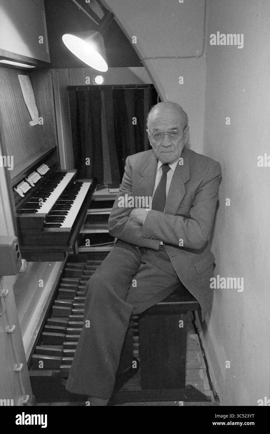 Mr. de Wilt Behind [the] Orgue, Orgues and Organists, 30-12-1985 Whizgle News, Dutch Desk, pays-Bas, 1950-2000 un homme âgé est assis avec soin dans une alcôve confortable à côté d'un orgue à pipe, ses bras croisés et une expression contemplative sur son visage. L'espace est intime, rehaussé par un éclairage doux qui attire l'attention sur les touches de l'orgue. Des textures riches et un sentiment de nostalgie emplissent la scène. Banque D'Images