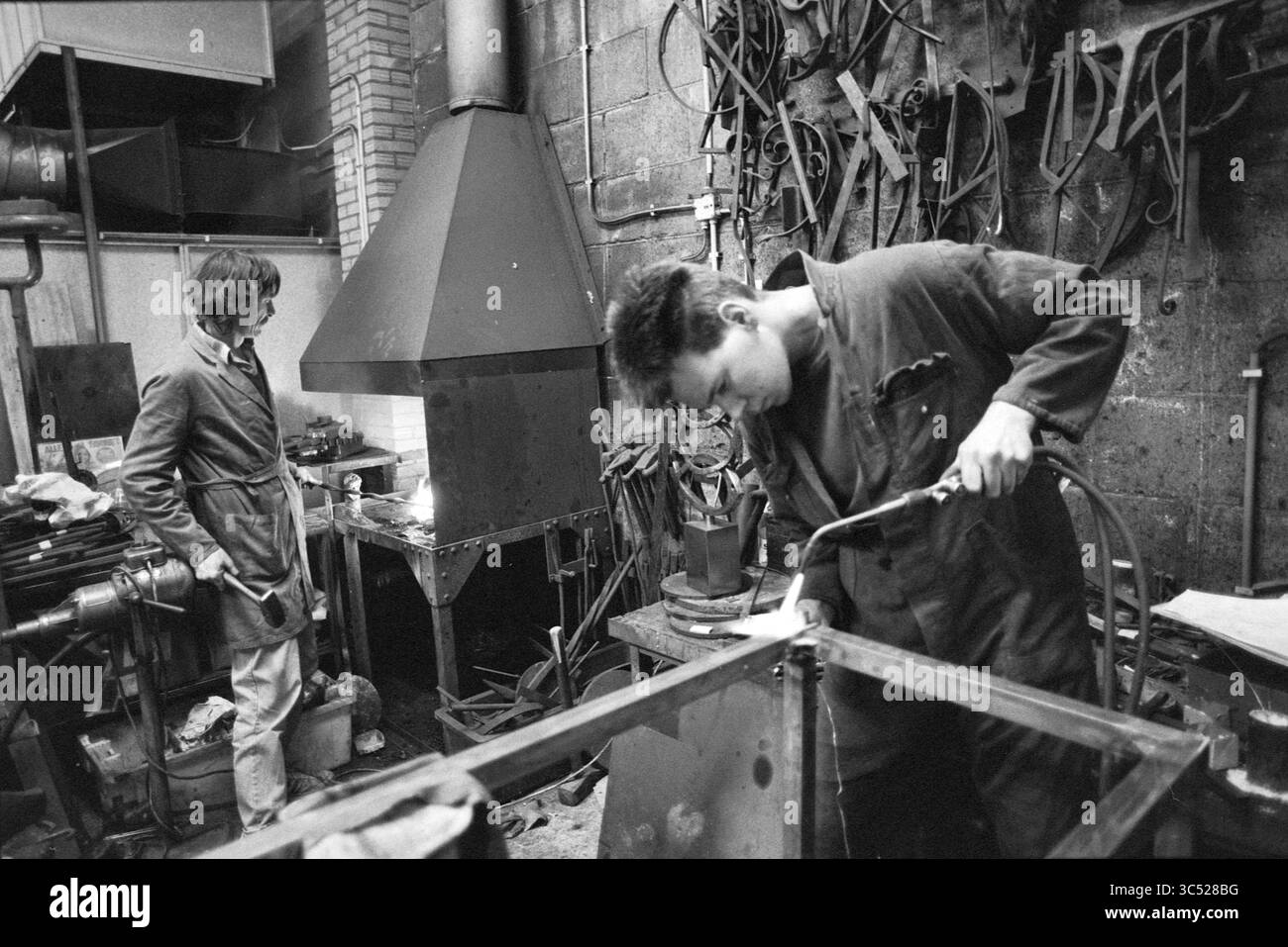 Atelier de soudage et de forgeage, 00-03-1989 Whizgle News, Dutch Desk, pays-Bas, 1950-2000 deux personnes travaillent dans un atelier rempli d'outils et d'équipements. L'un se concentre sur le soudage d'une pièce de métal, tandis que l'autre observe à proximité, vêtu d'une tenue de travail. L'environnement reflète une atmosphère industrielle, caractérisée par divers appareils métalliques et machines. Banque D'Images