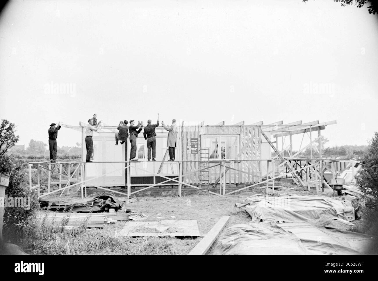 Bungalow en 2 jours à Heemskerk, construction, chantiers, 10-08-1960 Whizgle News, Dutch Desk, pays-Bas, 1950-2000 Un groupe de travailleurs collabore à la construction d'un bâtiment, avec des matériaux de levage et d'autres organisant la charpente. Des outils et des bâches dispersés sont visibles au premier plan, mettant l'accent sur l'environnement de travail actif. Banque D'Images