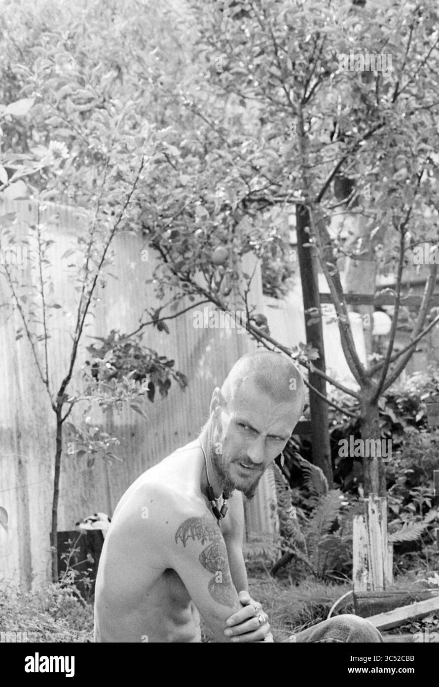 Scott Milller, artiste, H'lem, Haarlem, Nederland, 24-08-1994 Whizgle News, Dutch Desk, pays-Bas, 1950-2000 Un individu tatoué avec la tête rasée est assis dans un jardin, entouré d'arbres et de feuillage. La personne regarde en arrière avec une expression intense, tandis que la toile de fond présente une structure altérée. Banque D'Images