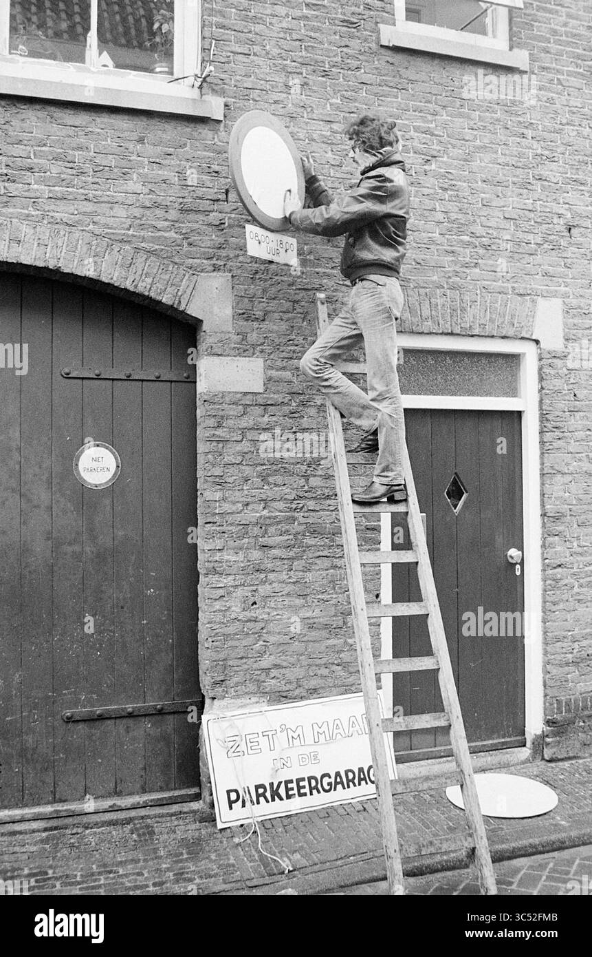 Action 'raccourci enfin levé' dans une rue étroite, 00-12-1974 Whizgle News, Dutch Desk, pays-Bas, 1950-2000 Un homme se balance sur une échelle, ajustant habilement un panneau circulaire monté sur un mur de briques, tandis que les panneaux ci-dessous indiquent les règles de stationnement. Deux portes flanquent la scène, ajoutant à l'atmosphère urbaine. Banque D'Images