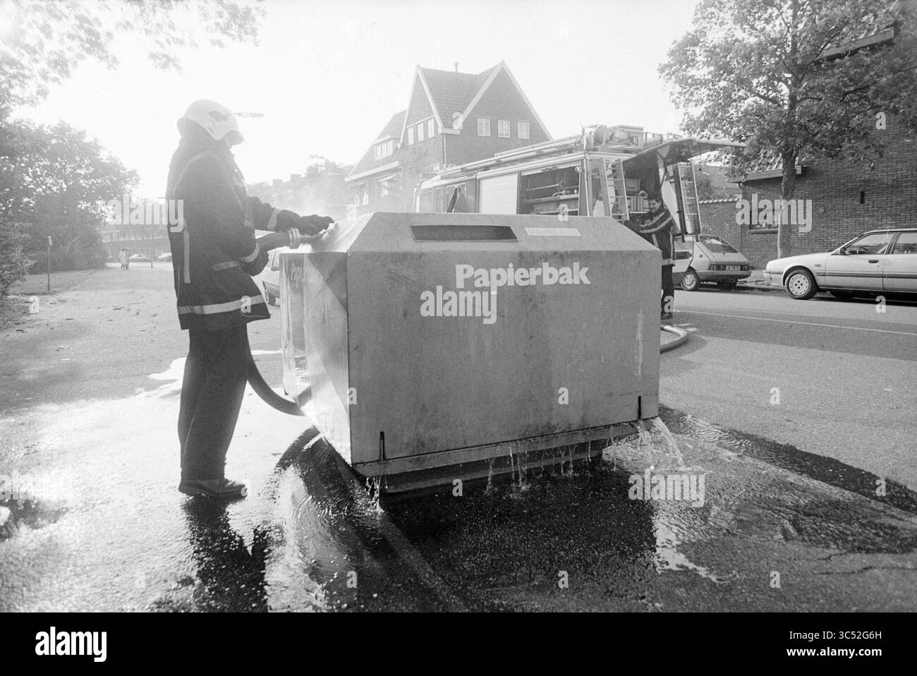 Poubelle en feu, 03-10-1992 Whizgle News, Dutch Desk, pays-Bas, 1950-2000 Un pompier utilise un tuyau pour nettoyer une grande poubelle de recyclage étiquetée « Papierbak », alors que l'eau coule dans la rue, soulignant les efforts déployés pour maintenir la propreté du public. En arrière-plan, un camion de pompiers et des voitures stationnées peuvent être vus. Banque D'Images