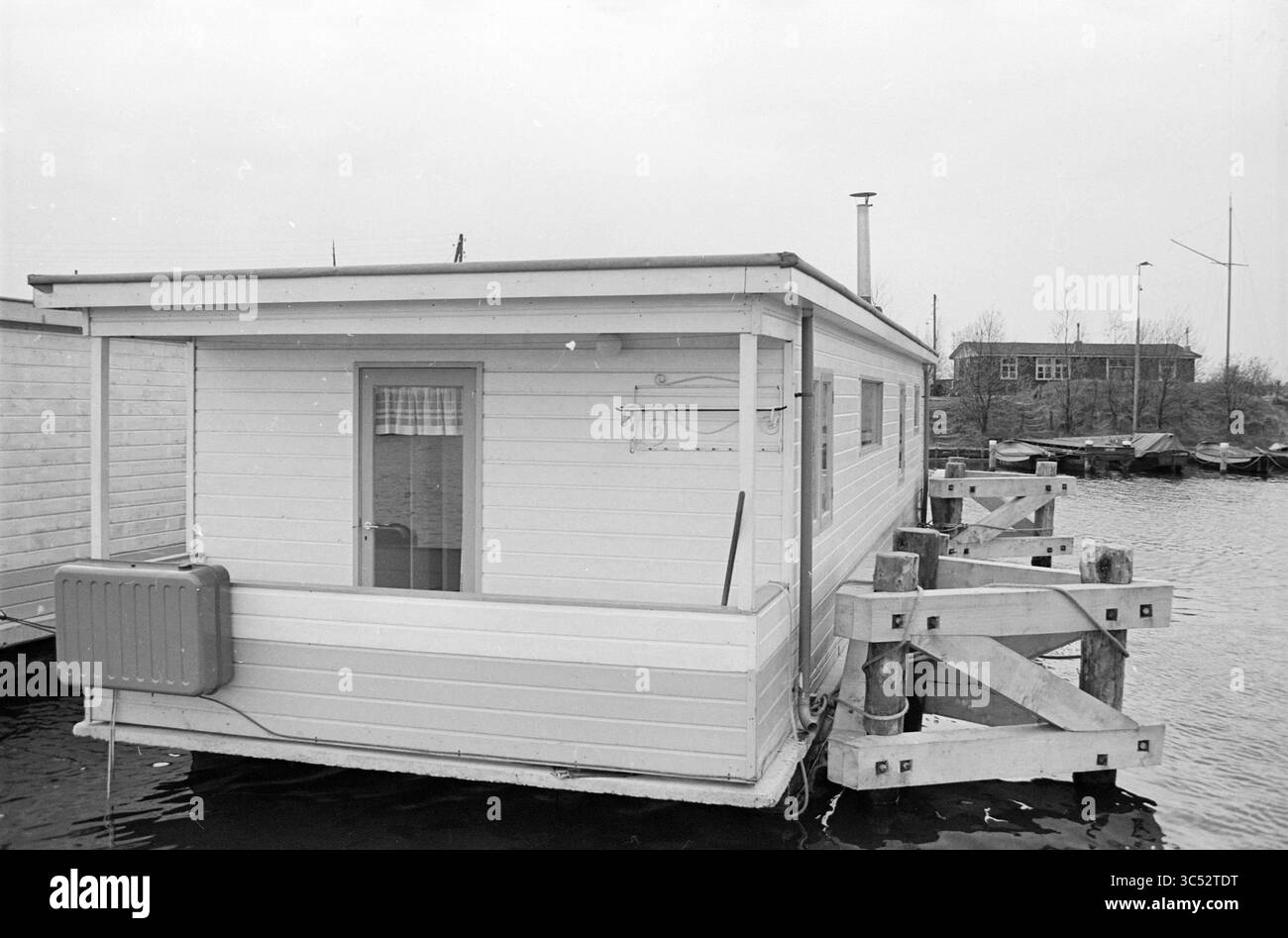 Remorquage de la péniche à travers le Ringvaart vers un nouvel emplacement, Aalsmeerderdijk Whizgle News, Dutch Desk, pays-Bas, 1950-2000 A small, la péniche en bois repose sur des pilotis au-dessus de l'eau, avec un design simple avec un porche avant et une cheminée. À proximité, des structures supplémentaires et des bateaux amarrés peuvent être vus le long du rivage, dans un ciel nuageux. Banque D'Images
