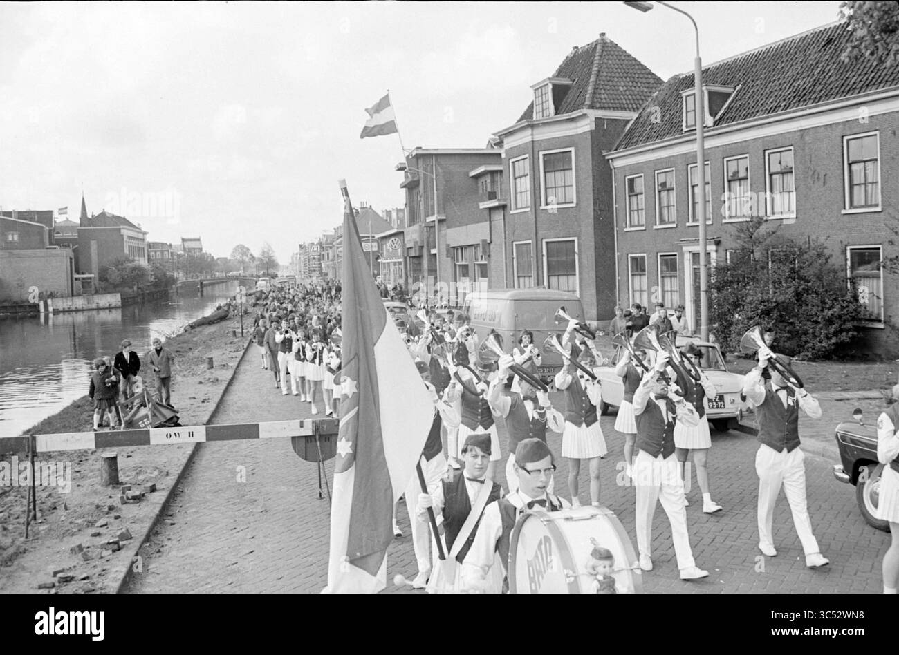 Défilé d'enfants sur patins à roulettes avec le groupe de musique Bato à Haarlem, Haarlem, pays-Bas, 00-00-1966 Whizgle News, Dutch Desk, pays-Bas, 1950-2000 Un défilé animé s'étend le long d'un canal, avec un fanion en uniforme blanc jouant des instruments. Les drapeaux branlent fièrement dans les airs alors qu'une foule se rassemble de chaque côté de la rue, certains spectateurs apparaissant engagés dans les festivités. Des bâtiments traditionnels bordent la route, ajoutant à l'atmosphère de célébration. Banque D'Images