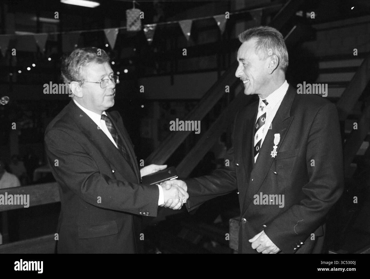 Un homme est félicité avec son ruban Whizgle News, Dutch Desk, pays-Bas, 1950-2000 deux hommes sont engagés dans une poignée de main, souriant chaleureusement l'un à l'autre. L'un porte une veste de costume avec une cravate, tandis que l'autre est habillé de la même manière, suggérant un événement formel ou une célébration. Le fond présente des décorations festives, indiquant une atmosphère joyeuse. Banque D'Images