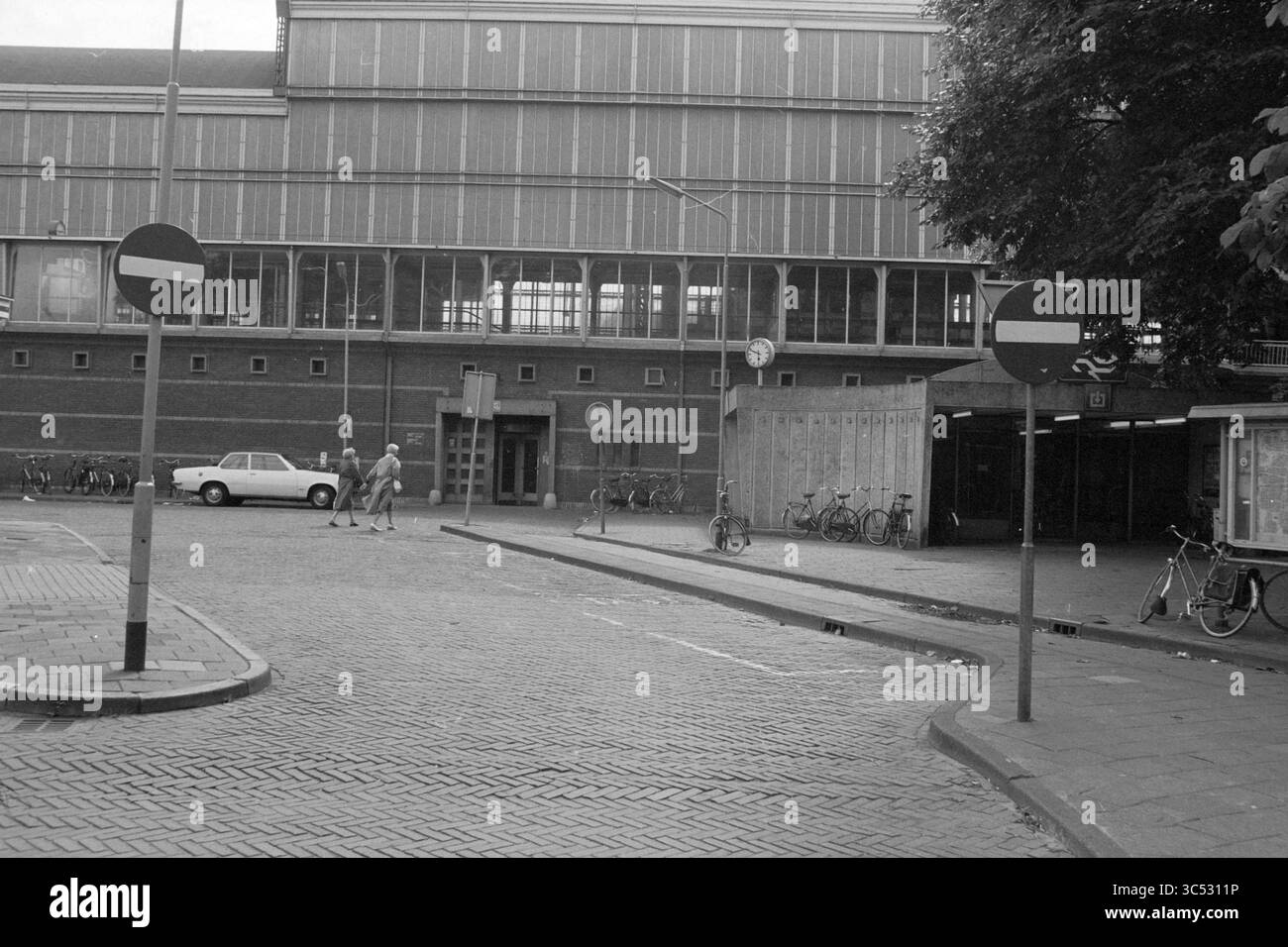 International et ext. Haarlem Station, Station, Haarlem, pays-Bas, 16-08-1981 Whizgle News, Dutch Desk, pays-Bas, 1950-2000 Une scène de rue présente un mélange de vélos et de véhicules garés près d'un grand bâtiment de style industriel. Deux piétons marchent vers l'entrée tandis que la signalisation indique les directions de la circulation. Des arbres bordent la zone, ajoutant une touche de verdure au milieu du cadre urbain. Banque D'Images