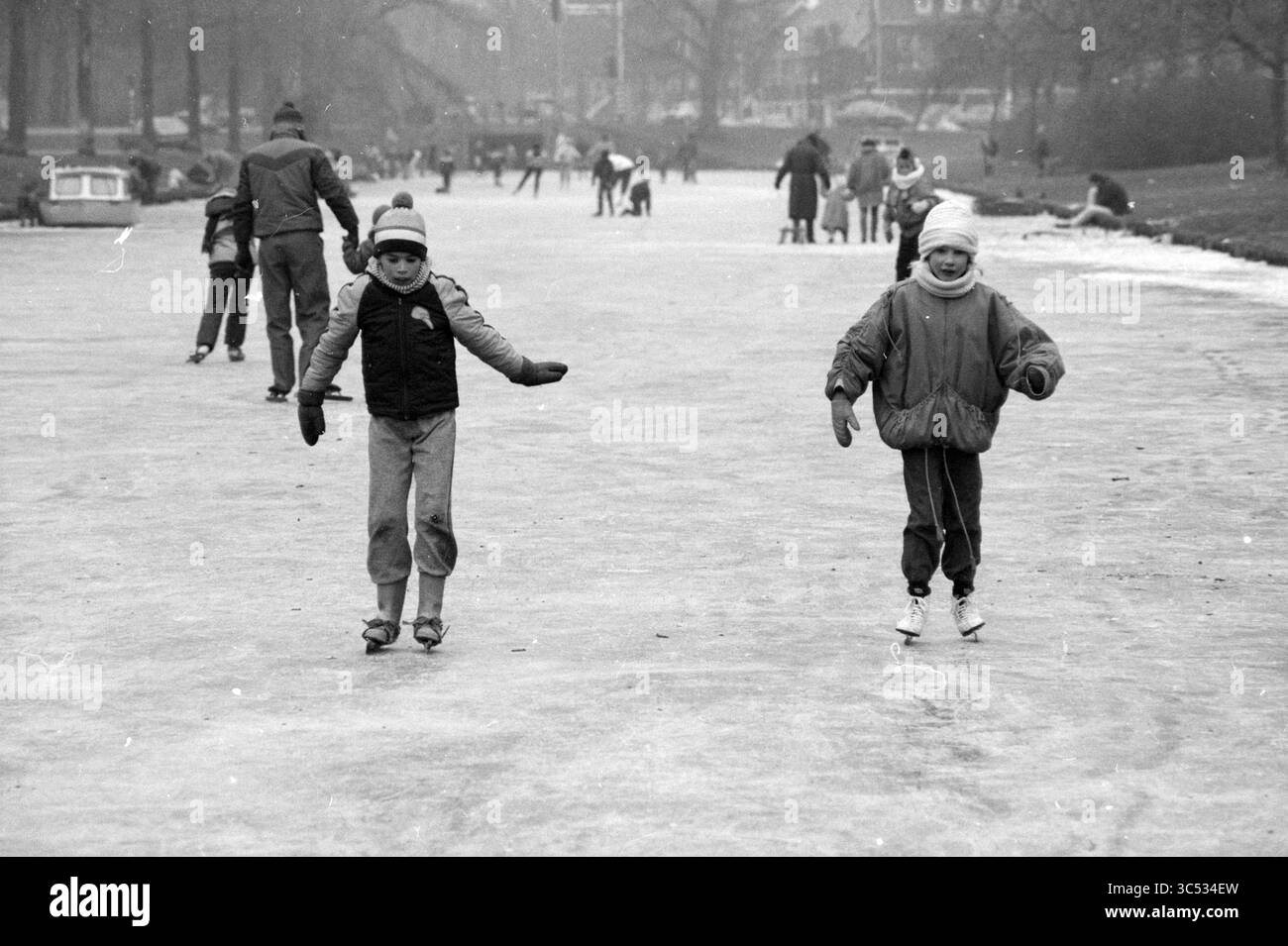 Patinage sur glace à Haarlem et autour, patinage sur glace, Haarlem, pays-Bas, 17-01-1987 Whizgle News, Dutch Desk, pays-Bas, 1950-2000 deux enfants glissent joyeusement sur une surface glacée, portant des vêtements chauds et des patins. La scène capture une atmosphère animée avec d'autres patineurs en arrière-plan, mettant en valeur une communauté profitant d'une journée d'hiver. Banque D'Images