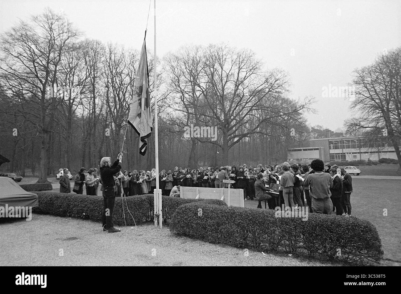 Actions chez LES DSI formation sportive chez Duinlust Whizgle News, Dutch Desk, pays-Bas, 1950-2000 Un groupe de personnes se réunit à l'extérieur près d'un mât de drapeau, alors qu'un individu lève le drapeau. Les arbres bordent le fond, et la scène transmet un sentiment de solennité et de rassemblement communautaire. Banque D'Images