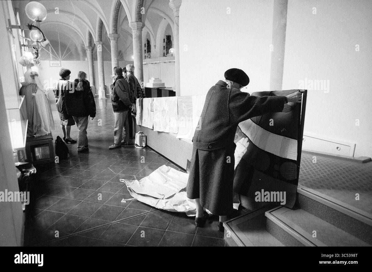 Installation du réfectoire d'exposition, 23-02-1992 Whizgle News, Dutch Desk, pays-Bas, 1950-2000 Une personne dans un béret examine et ajuste des panneaux de tissu dans une galerie spacieuse, tandis qu'un petit groupe de personnes conversent à proximité, entouré de détails architecturaux et d'un éclairage tamisé. Banque D'Images