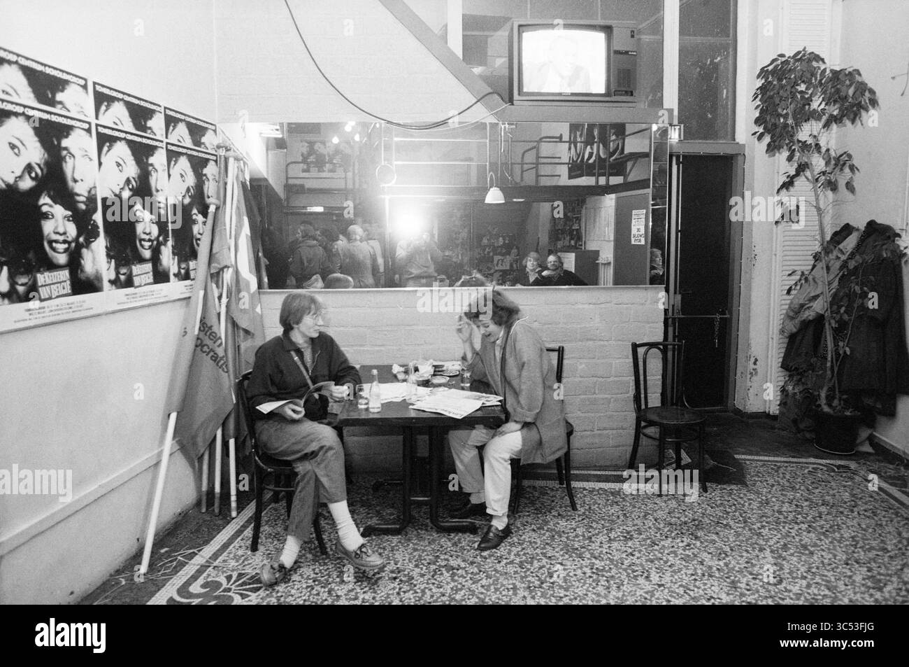 Café politique dans le Toneelschuur, café politique, Haarlem, pays-Bas, 11-03-1986 Whizgle News, Dutch Desk, pays-Bas, 1950-2000 deux personnes assises à une petite table dans un espace animé, engagées dans la conversation autour d'un verre. Derrière eux, un grand miroir reflète l'atmosphère animée remplie de gens et d'affiches vintage sur les murs. Un téléviseur affiche des images, ajoutant à l'environnement dynamique. Banque D'Images