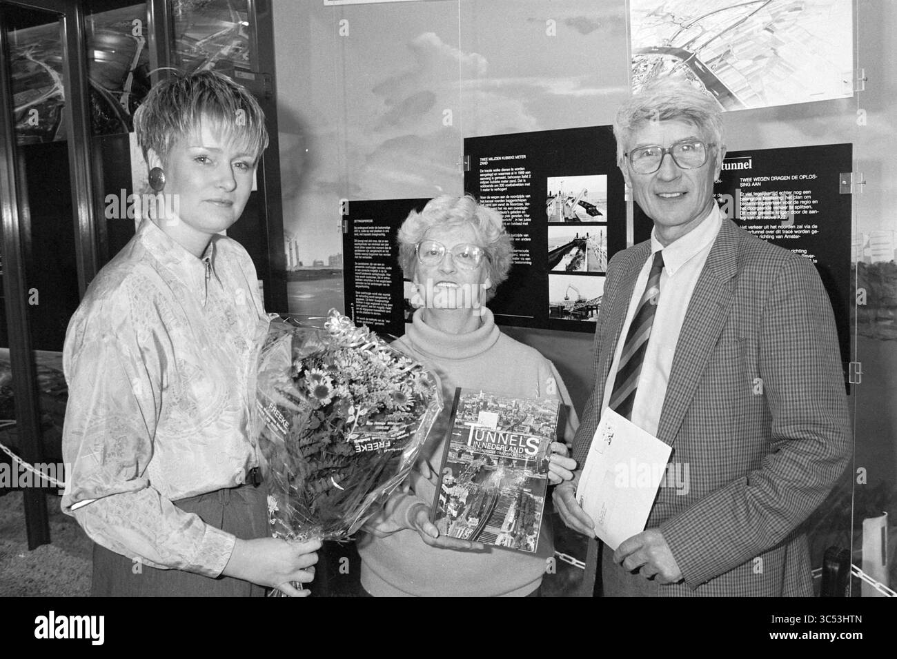 10,000ème visiteur du centre d'information Wijkertunnel, 19-04-1994 Whizgle News, Dutch Desk, pays-Bas, 1950-2000 trois personnes se tiennent ensemble, souriant dans un cadre d'affichage. Une personne tient un bouquet de fleurs, tandis qu'une autre présente un livre intitulé 'TUNNELS'. Le groupe semble engagé et célébrant, commémorant probablement un événement spécial ou une réalisation liée au sujet du livre. Des panneaux informatifs sont visibles en arrière-plan, ajoutant du contexte à l'occasion. Banque D'Images