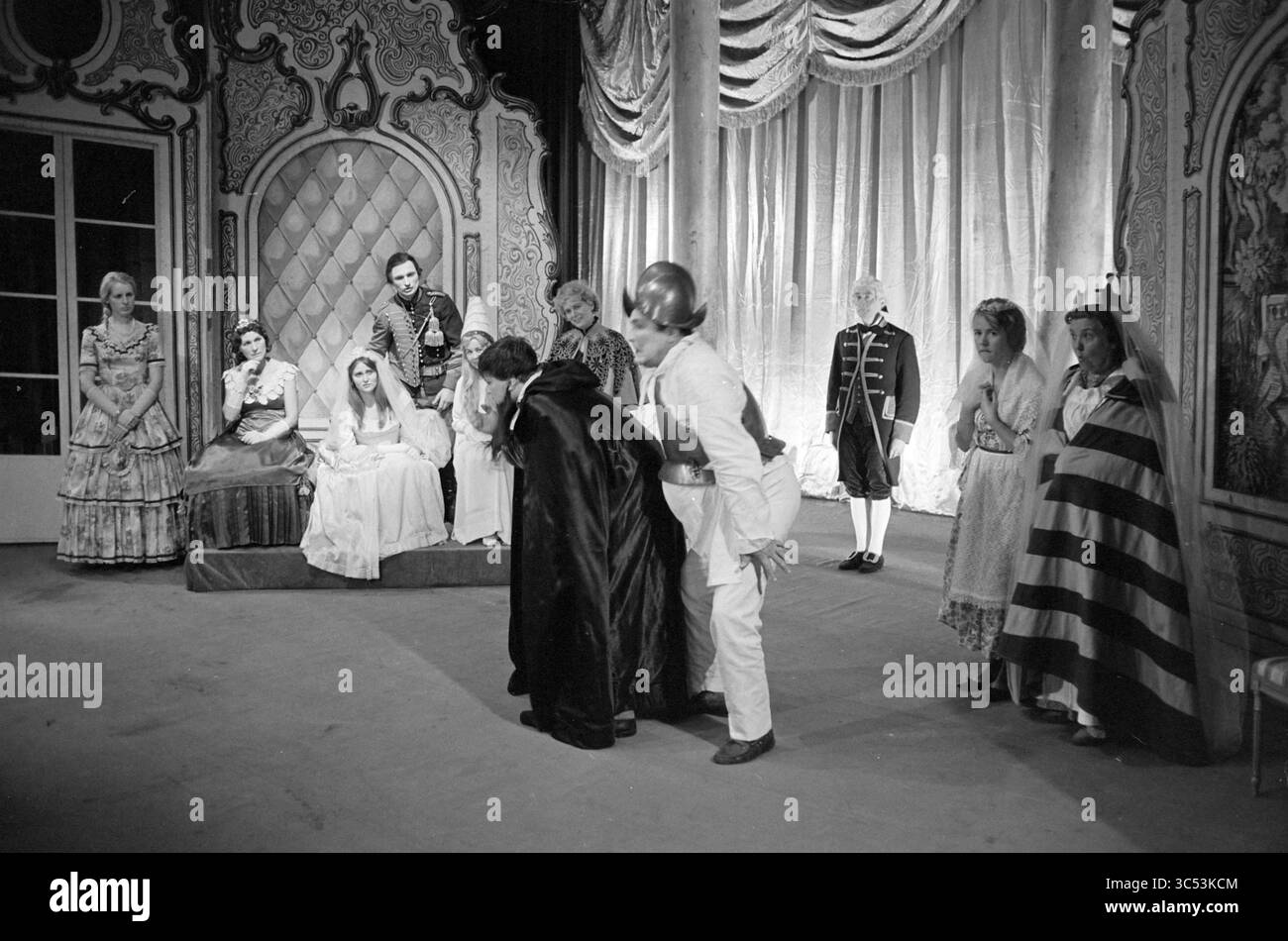 Théâtre, drame costumé, comédie Whizgle News, Dutch Desk, pays-Bas, 1950-2000 Un groupe d'acteurs vêtus de costumes élaborés est rassemblé sur scène, certains debout et d'autres assis. La scène semble représenter un moment dramatique, mettant en valeur des expressions d'intrigue et de tension parmi les personnages. L'arrière-plan et le mobilier ornés suggèrent un décor théâtral, tandis que divers costumes évoquent un thème historique ou fantastique. Banque D'Images