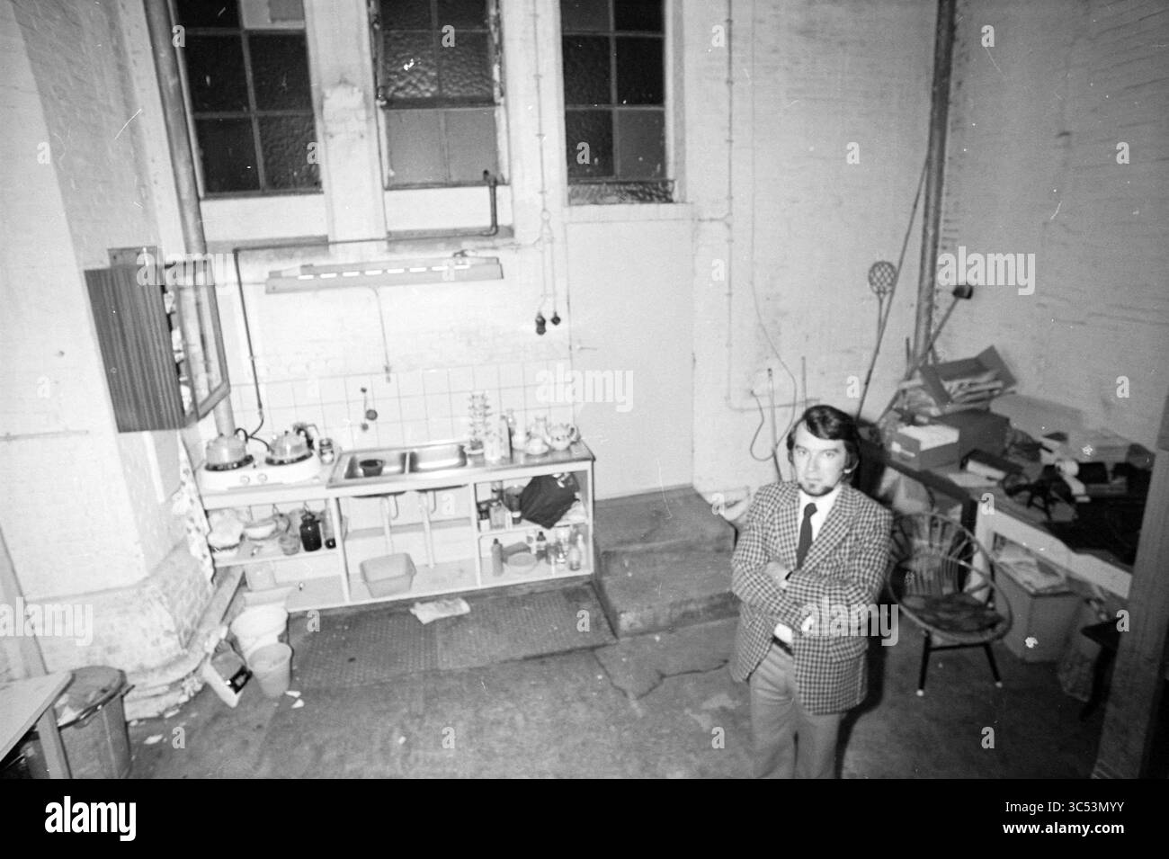 M.A.I. Office Mr. Gorrits., social Work, 07-08-1974 Whizgle News, Dutch Desk, pays-Bas, 1950-2000 Un homme se tient confiant, les bras croisés, dans un espace de cuisine de style industriel peu meublé. La chambre dispose d'un coin lavabo avec vaisselle et ustensiles, tandis que les environs sont encombrés de divers articles, ajoutant à l'atmosphère éclectique. De grandes fenêtres apportent la lumière naturelle, éclairant les murs usés. Banque D'Images
