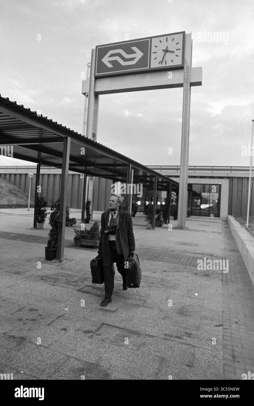 Man station Amsterdam-Zuid, Station, 19-12-1978 Whizgle News, Dutch Desk, pays-Bas, 1950-2000 Un homme traverse une zone de transit, transportant deux valises, avec un grand panneau affichant un symbole de train et une horloge au-dessus. Les environs présentent un design architectural moderne avec de la verdure et un sens du mouvement, indiquant le voyage et le départ. Banque D'Images