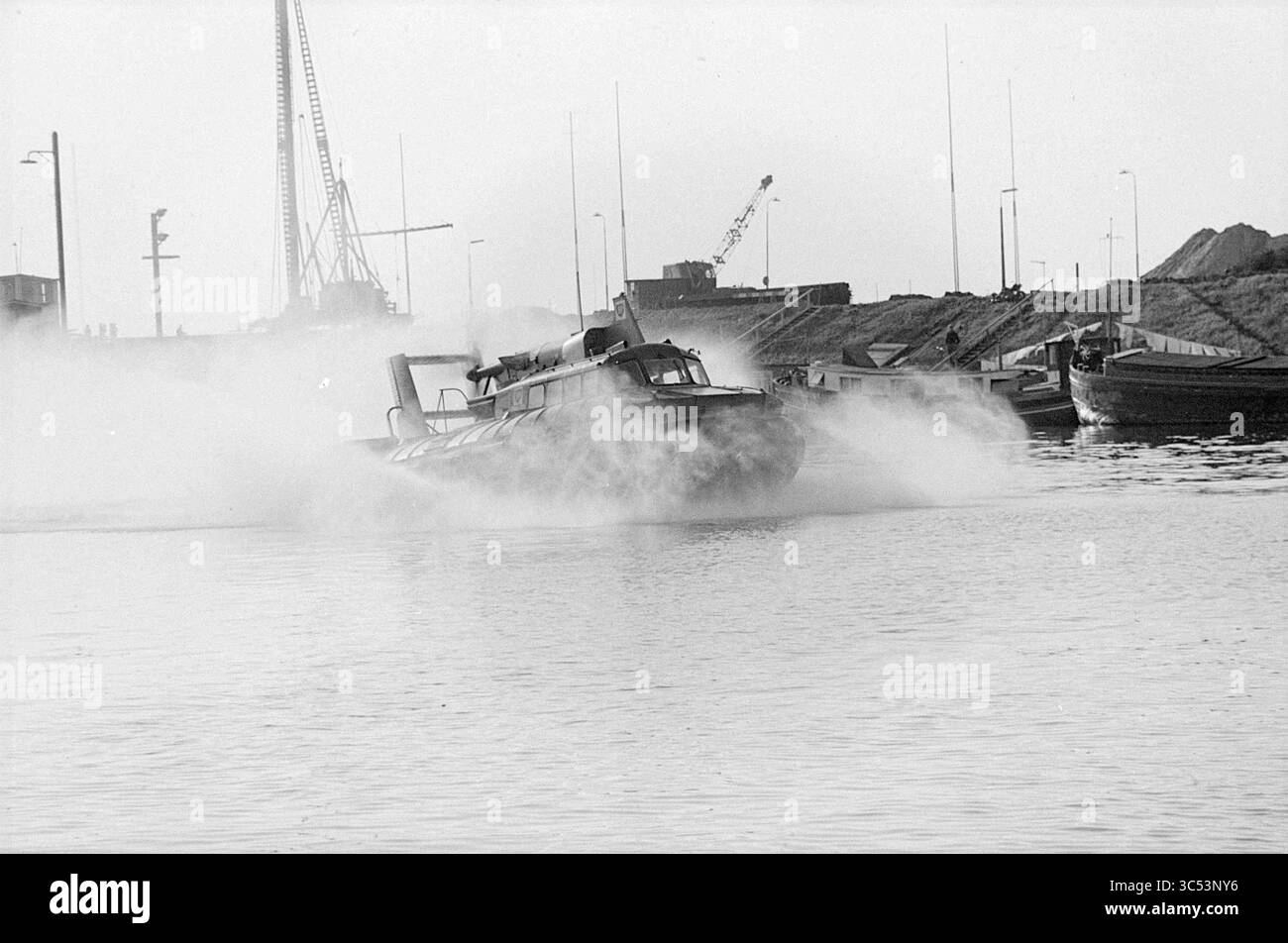 Aéroglisseur, navires, 11-05-1965 Whizgle News, Dutch Desk, pays-Bas, 1950-2000 Un bateau rapide coupe l'eau, envoyant une brume lorsqu'il navigue près d'un quai animé, où grues et bateaux sont visibles en arrière-plan. Banque D'Images