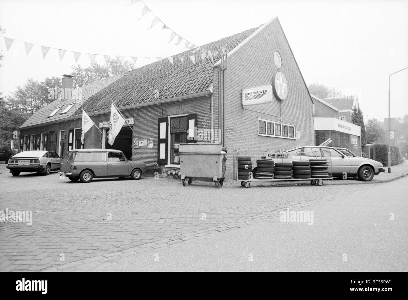 Garage Sud (extérieur), Haarlem, Kleine Houtweg, pays-Bas, 00-00-1986 Whizgle News, Dutch Desk, pays-Bas, 1950-2000 Un magasin automobile pittoresque dispose d'un bâtiment vintage orné de drapeaux et de panneaux. Deux voitures classiques sont garées devant, tandis qu'une pile de pneus et une benne à ordures se trouvent à proximité, ajoutant au charme rustique de la scène. Banque D'Images