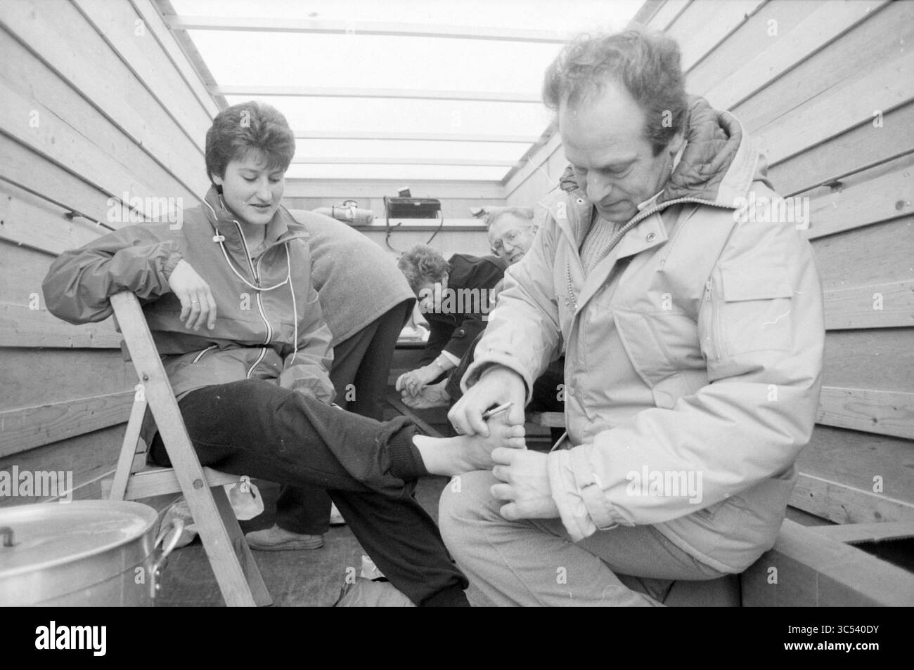 Winter Walk Courier, Walking, 20-02-1988 Whizgle News, Dutch Desk, pays-Bas, 1950-2000 Un homme tend le pied d'une femme pendant qu'elle se détend en position assise, portant des écouteurs. En arrière-plan, deux autres sont engagés dans une activité, créant une atmosphère décontractée et connectée. Des murs en bois et un cadre simple soulignent un sentiment d'intimité et de soin. Banque D'Images