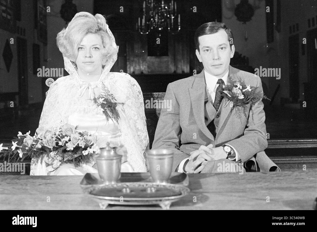 Mariage Warnars - van de Wel, mariage, 26-11-1969 Whizgle News, Dutch Desk, pays-Bas, 1950-2000 Une mariée et un marié s'assoient côte à côte à une table de cérémonie, tous deux habillés formellement. La mariée porte une robe blanche avec un voile de dentelle et tient un bouquet de fleurs, tandis que le marié est dans un costume orné d'un corsage de fleurs. Leurs expressions sont sérieuses alors qu'elles se préparent pour la cérémonie à venir, avec un intérieur faiblement éclairé avec des détails décoratifs en arrière-plan. Banque D'Images