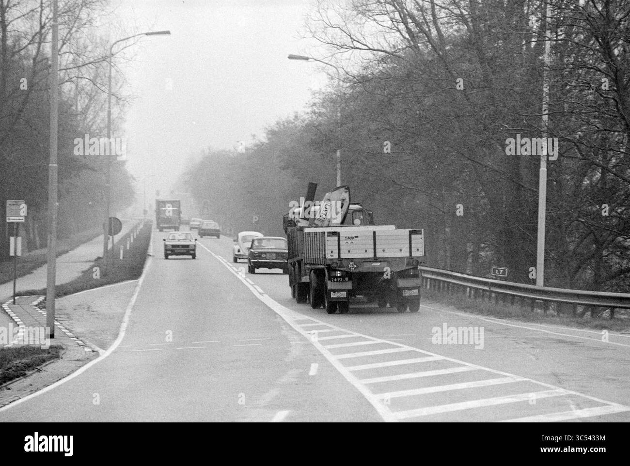 Kruisweg (S21) entre Cruquiusweg et IJweg, circulation, sécurité routière, routes, construction de routes, plantation de routes, Haarlemmermeer, Kruisweg, 22-12-1980 Whizgle News, Dutch Desk, pays-Bas, 1950-2000 Une scène de route brumeuse présente des véhicules, y compris un camion chargé de matériaux, naviguant le long d'une route divisée bordée d'arbres. Les panneaux de signalisation et les lampadaires sont visibles, laissant entendre une journée calme et nuageuse. Banque D'Images