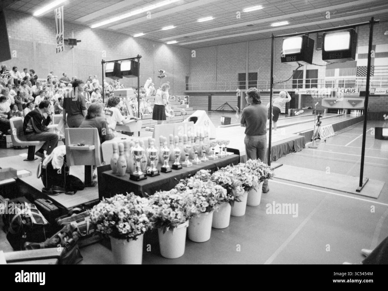 Finales de bowling Beyneshal, Bowling, Haarlem, pays-Bas, 21-06-1987 Whizgle News, Dutch Desk, pays-Bas, 1950-2000 un événement intérieur animé mettant en vedette une foule réunie dans des gradins, avec des participants engagés dans une compétition de bowling. Au premier plan, un ensemble de trophées ornés de fleurs évoque l'importance de la journée, tandis qu'un tableau de bord et des écrans de télévision capturent l'action sur les voies. Banque D'Images