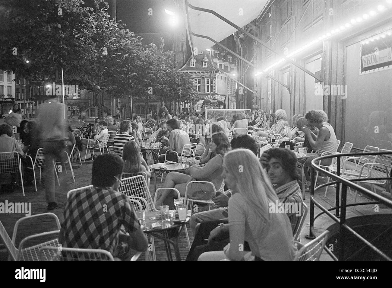 Terrasse Brinkmann au soir d'été, terrasse, photos d'été, Haarlem, Grote Markt, pays-Bas, 08-08-1975 Whizgle News, Dutch Desk, pays-Bas, 1950-2000 Une scène de café en plein air animée, remplie de gens qui apprécient la conversation et les boissons à des tables sous des auvents ombragés. Les arbres et les lumières décoratives ajoutent à l'atmosphère, tandis qu'un mélange de tenues décontractées reflète un rassemblement social détendu. Banque D'Images