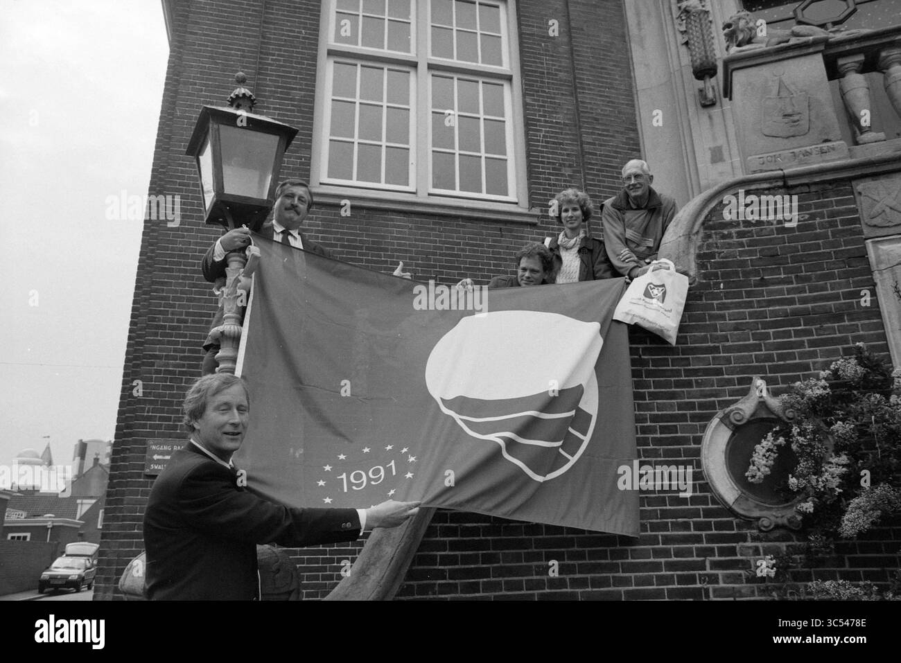 Drapeau écologique pour Zandvoort, Zandvoort, 06-06-1991 Whizgle News, Dutch Desk, pays-Bas, 1950-2000 Un groupe de cinq personnes arbore fièrement un grand drapeau orné d'une coquille stylisée, commémorant l'année 1991. Ils sont positionnés devant un bâtiment en briques avec des éléments architecturaux décoratifs, et certains se tiennent debout sur un rebord tandis que d'autres s'appuient contre le mur. Banque D'Images
