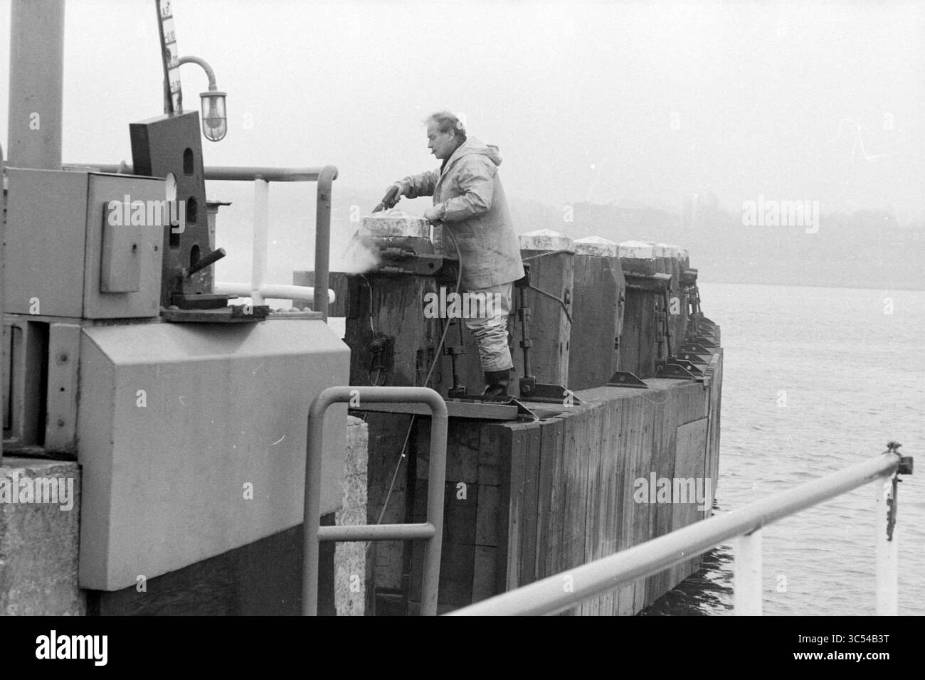 Freins de ferry nettoyage ferry Velsen, ponts, nettoyage, Velsen, 13-03-1985 Whizgle News, Dutch Desk, pays-Bas, 1950-2000 Un ouvrier se tient sur un quai, appliquant soigneusement une couche de peinture sur une barrière tout en étant entouré par un paysage serein de front de mer. L'atmosphère est calme, avec une légère brume planant dans l'air. Banque D'Images