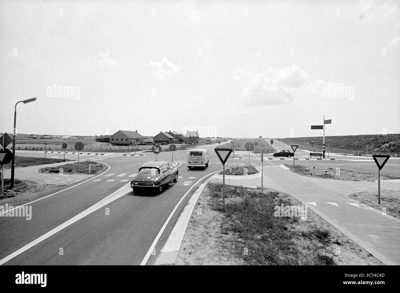 Crossing de Zilk, routes, construction de routes, plantation de routes, 29-06-1972 Whizgle News, Dutch Desk, pays-Bas, 1950-2000 Une scène vintage capture un rond-point avec des véhicules naviguant à l'intersection circulaire, entouré d'un paysage clairsemé et de quelques bâtiments au loin. Les panneaux routiers indiquent la direction et les points d'écoulement, tandis que les nuages dérivent au-dessus dans un ciel dégagé. Banque D'Images