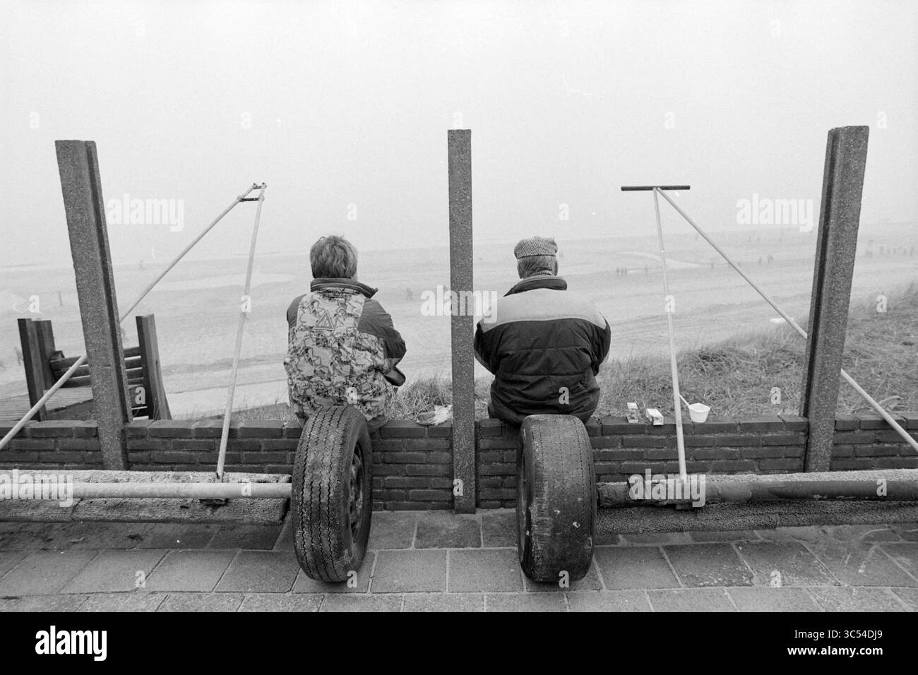 Winter Walk, 13-02-1993 Whizgle News, Dutch Desk, pays-Bas, 1950-2000 deux individus assis sur des pneus, regardant vers une plage lointaine. Ils sont entourés d'une clôture rustique qui encadre leur vue, créant un sentiment de solitude et de réflexion. L’ambiance est calme, évoquant un moment de contemplation au bord de la mer. Banque D'Images