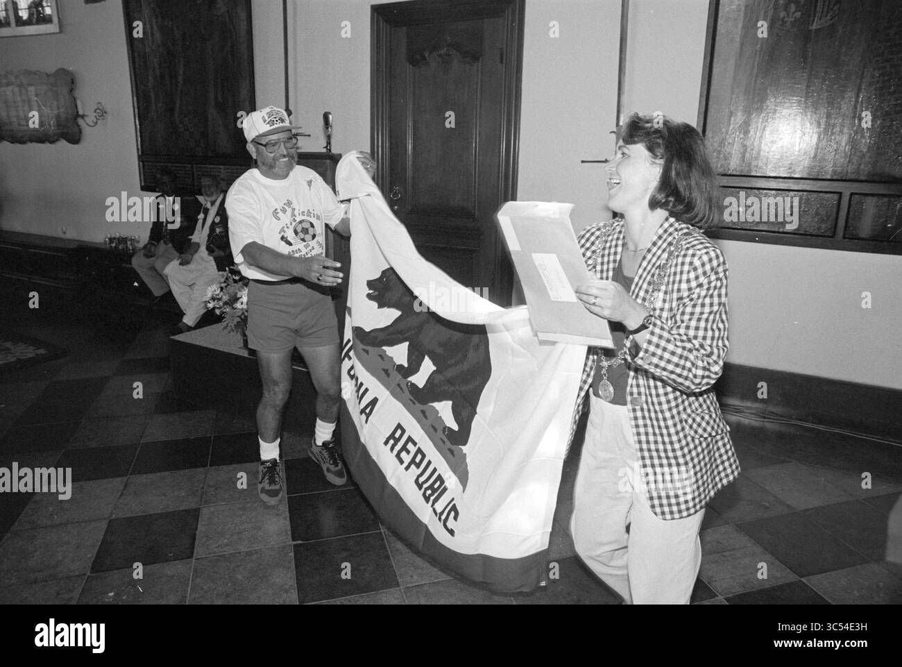 Montrer le drapeau de la République de Californie, 02-08-1994 Whizgle News, Dutch Desk, pays-Bas, 1950-2000 Un homme en tenue décontractée présente un drapeau avec un motif ours à une femme en blazer à carreaux, qui sourit en le recevant. Le paramètre semble être à l'intérieur, avec des détails ornés en arrière-plan. Banque D'Images