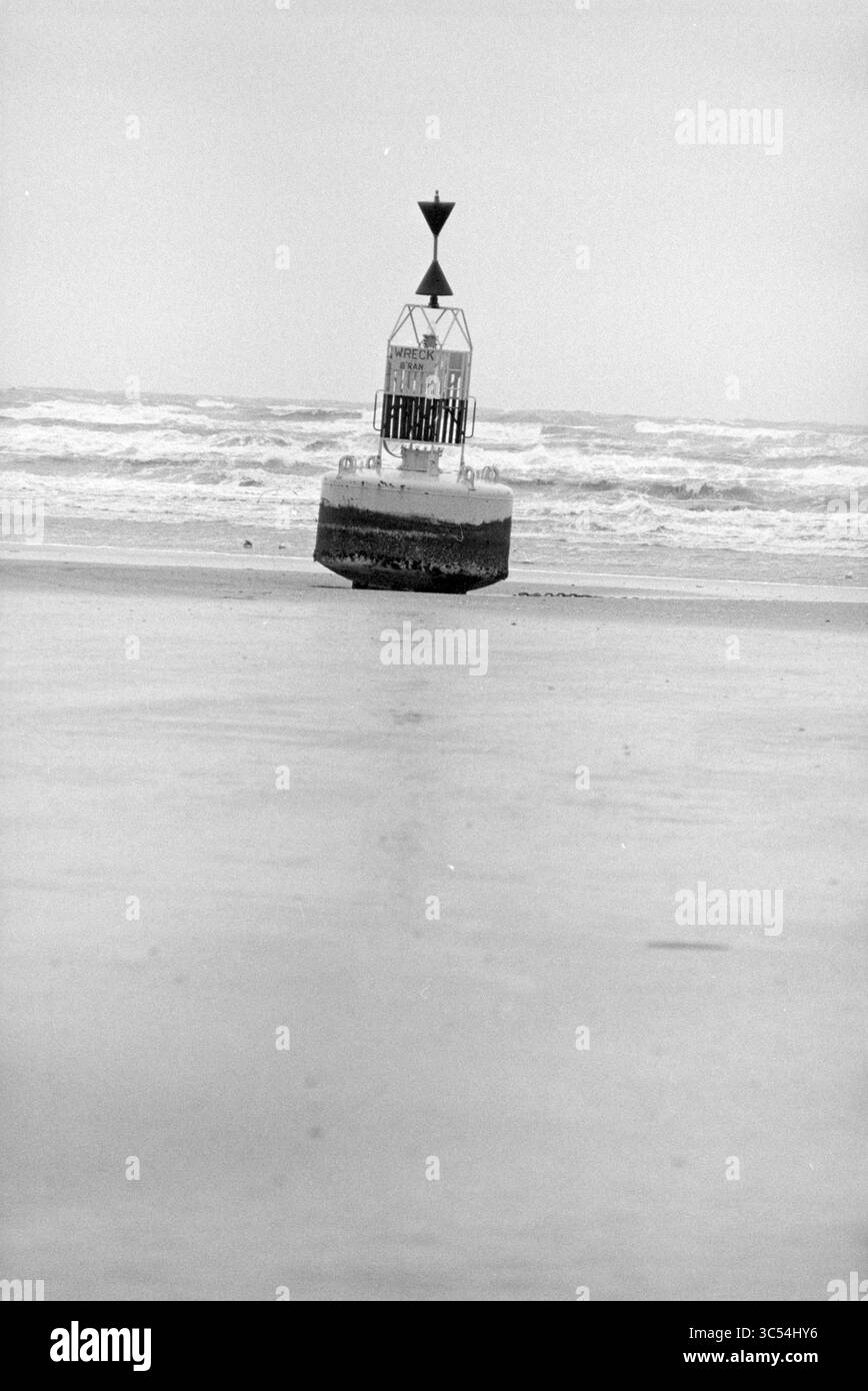 Bouée lavée IJmuiden, IJmuiden, pays-Bas, 25-01-1993 Whizgle News, Dutch Desk, pays-Bas, 1950-2000 Une bouée solitaire se dresse au bord de l'eau, entourée de douces vagues sous un ciel couvert. Sa forme distinctive et ses marques créent un point focal frappant contre le rivage tranquille. Banque D'Images