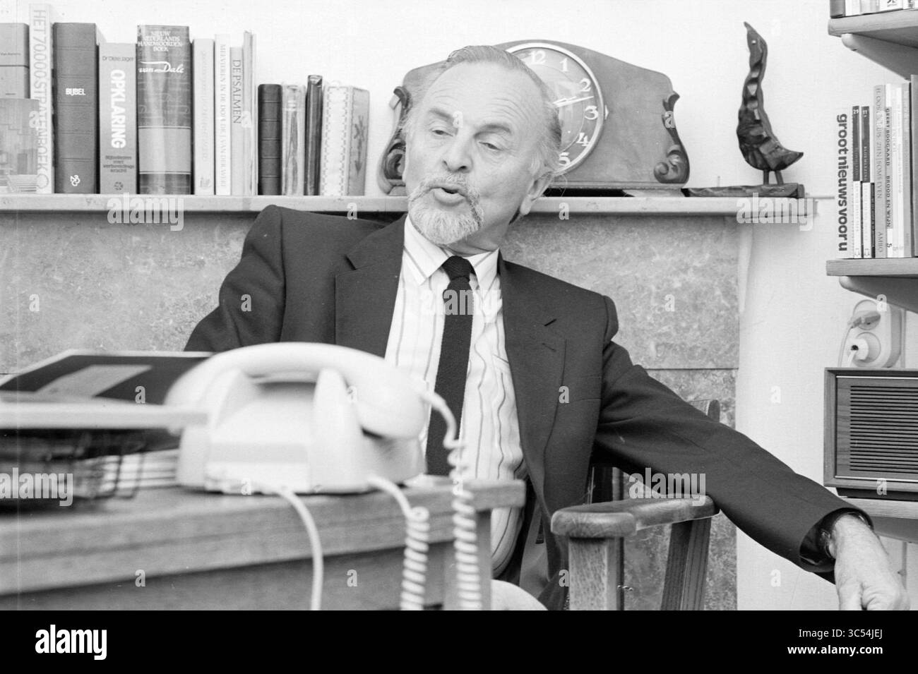 Pastor Timmermans, IJmuiden, Pastor, IJmuiden, pays-Bas, 19-06-1985 Whizgle News, Dutch Desk, pays-Bas, 1950-2000 un homme âgé en costume est assis confortablement à un bureau, engagé dans la conversation, avec des livres bordant une étagère voisine et une horloge vintage en arrière-plan. Un téléphone repose sur le bureau, ajoutant à l'atmosphère nostalgique. Banque D'Images