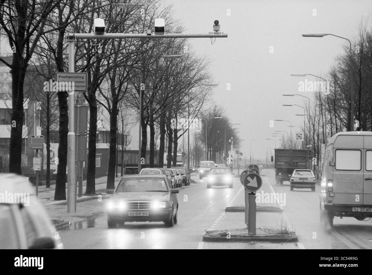 Caméra de circulation, Oudeweg, Haarlem, Oudeweg, pays-Bas, 02-12-1996 Whizgle News, Dutch Desk, pays-Bas, 1950-2000 Une scène de rue animée avec des véhicules naviguant sur une route baignée de pluie, flanquée d'arbres bordant les côtés. Une caméra de circulation est montée sur un poteau, surveillant le flux des voitures lorsqu'elles approchent d'une intersection circulaire en avant. Les panneaux de signalisation donnent des instructions aux conducteurs, tandis que l'atmosphère silencieuse suggère une journée calme et nuageuse. Banque D'Images