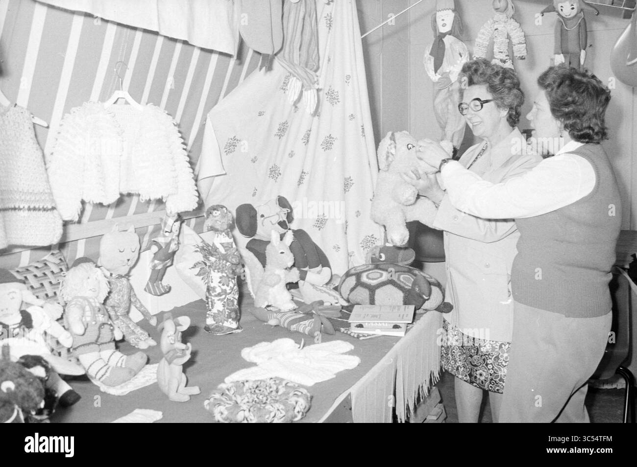 Stalls in Community Center., Stalls, 25-05-1974 Whizgle News, Dutch Desk, pays-Bas, 1950-2000 deux femmes sont entourées d'un éventail de jouets et d'objets artisanaux faits à la main, y compris des animaux en peluche et des poupées, tandis qu'une femme tient un jouet en peluche. Le cadre respire un sens de la créativité et de la communauté, mettant en valeur l'artisanat artisanal. Banque D'Images
