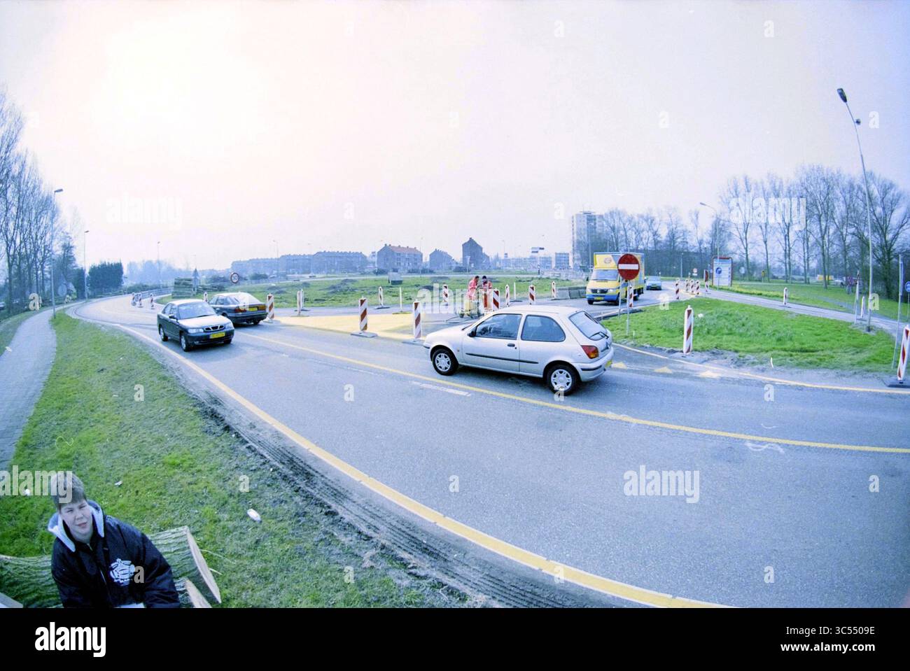 Delftplein Roundabout, Haarlem, Delftplein, Nederland, 27-03-2001 Whizgle News, Dutch Desk, pays-Bas, 1950-2000 Un rond-point animé entouré de travaux, avec des voitures naviguant sur la route circulaire. Au premier plan, une personne est assise sur un banc, observant l'activité, tandis que les piétons traversent à proximité au milieu des panneaux de signalisation indiquant des déviations. La scène capture un mélange de vie urbaine et de développement continu. Banque D'Images