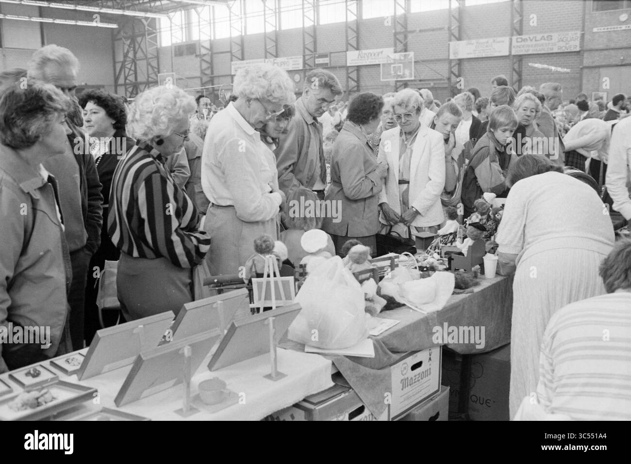 Foire artisanale Whizgle News, Dutch Desk, pays-Bas, 1950-2000 Une scène de marché intérieur animée remplie d'une foule de personnes parcourant divers stands et expositions, mettant en vedette l'artisanat et les marchandises faites à la main. Les acheteurs enthousiastes examinent de près les articles sur les tables, tandis que les conversations et l'excitation remplissent l'air. Banque D'Images